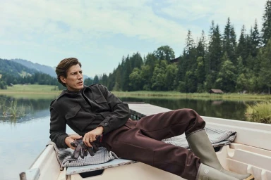 Model Oriol Elcacho reclining in a white rowboat on a mountain lake wearing a dark jacket, brown trousers, and rubber boots.