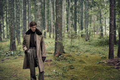 Model Oriol Elcacho stands in a dense pine forest wearing a checked houndstooth overcoat, turtleneck, and tweed trousers.