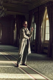 Oriol Elcacho in a long taupe trench coat and pleated trousers standing beside tall draped windows in a grand sunlit hall.