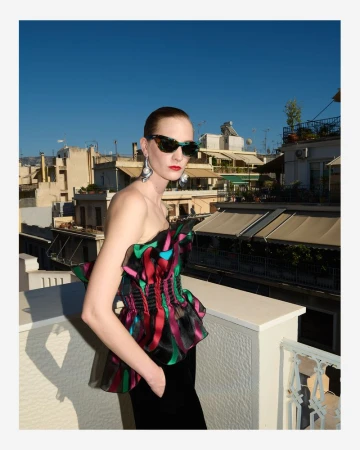 Fashion shot of a woman wearing sunglasses, a ruffled strapless top and dark pants on a rooftop with city buildings in the background.