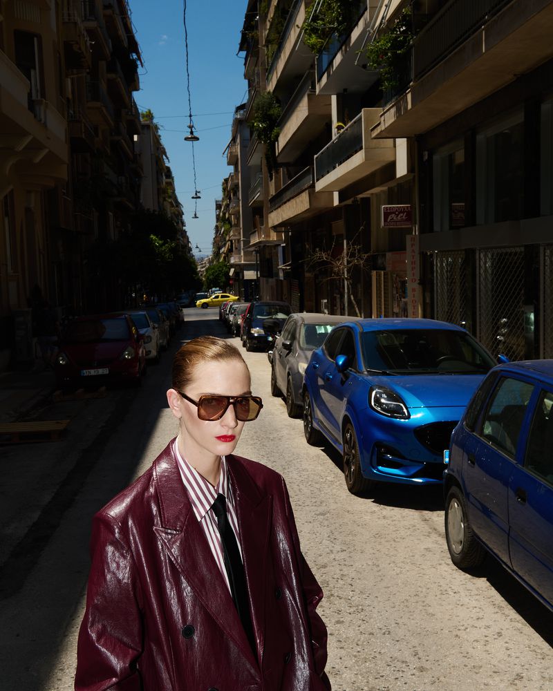 Fashionable woman in a maroon jacket poses on a city street lined with parked cars, showcasing an urban, editorial style.