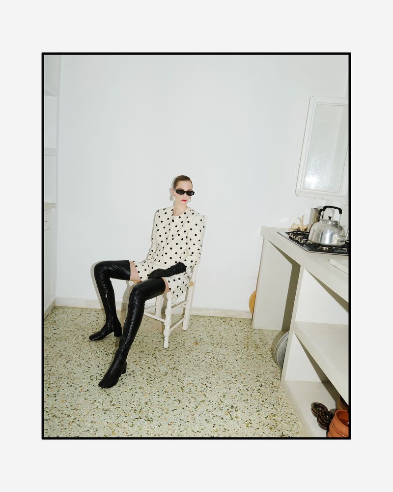 A fashionable woman sits in a kitchen wearing a black and white polka dot dress and shiny black thigh-high boots with sunglasses.