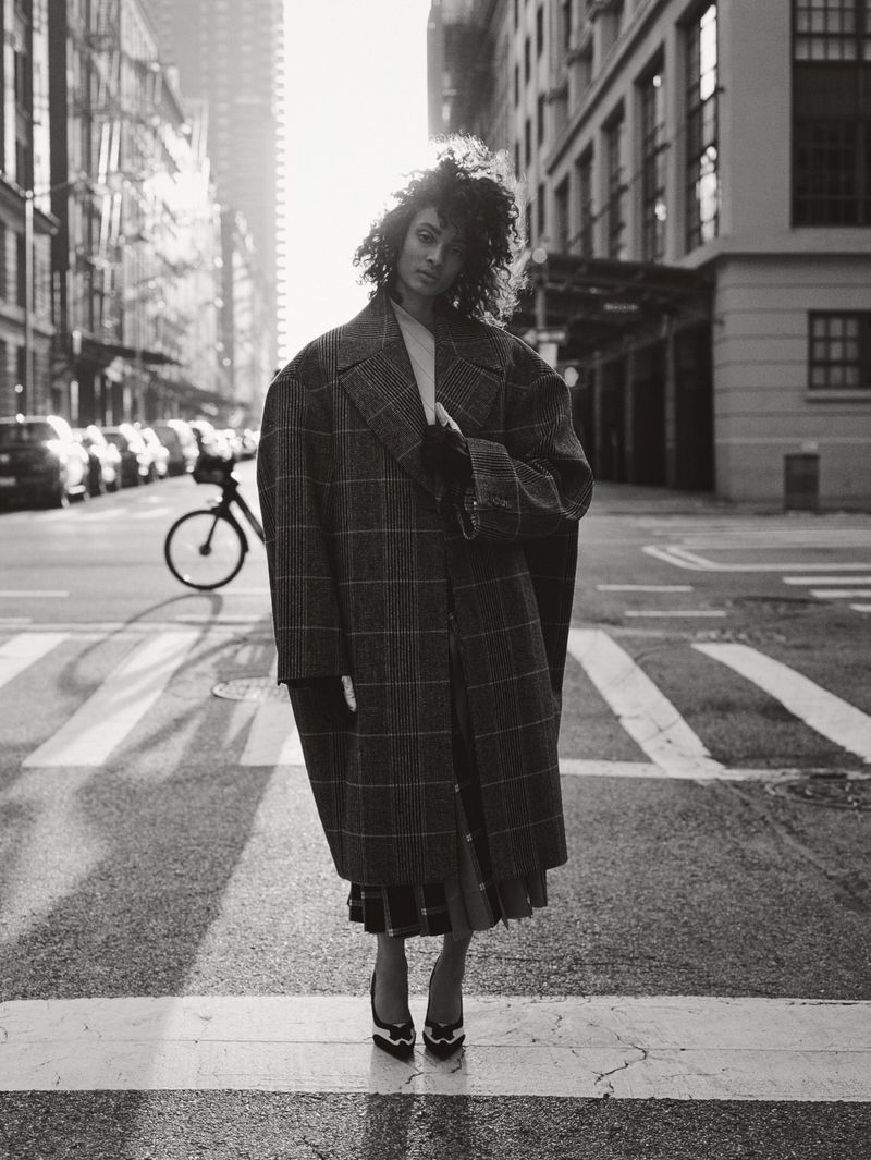 Monochrome portrait of a woman in a plaid coat standing on a crosswalk in a city, with tall buildings in the background on a sunny day.