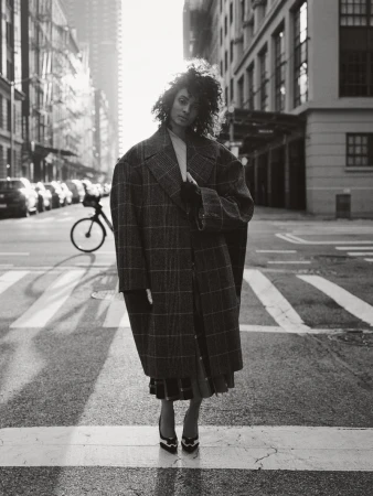 Monochrome portrait of a woman in a plaid coat standing on a crosswalk in a city, with tall buildings in the background on a sunny day.
