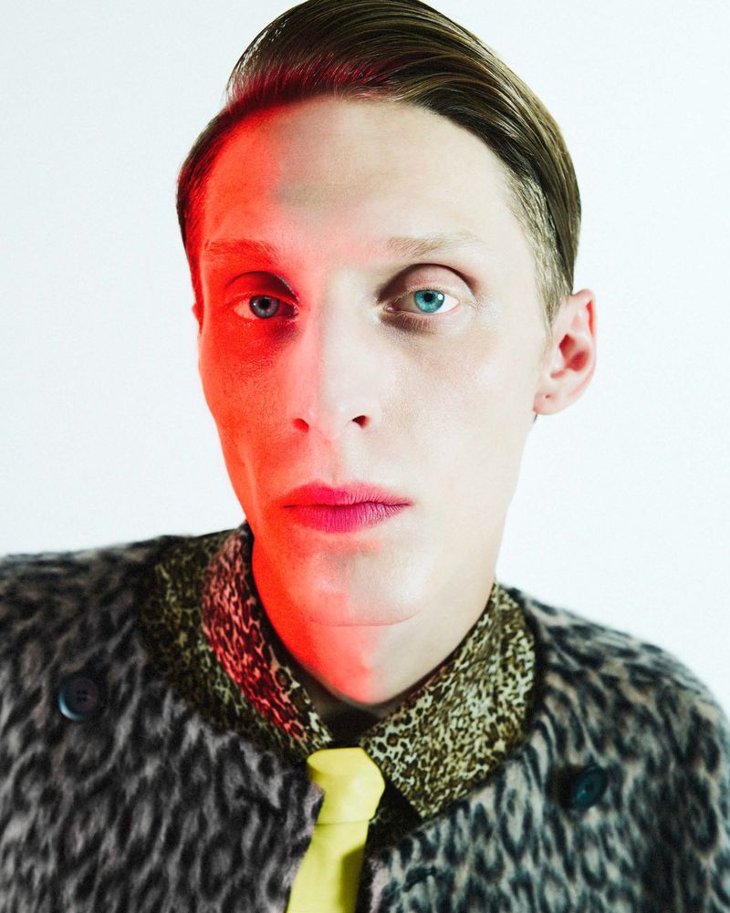 Portrait of a fair-skinned adult male with slicked-back brown hair, wearing a patterned collared shirt, animal print jacket, and yellow tie, red lighting.
