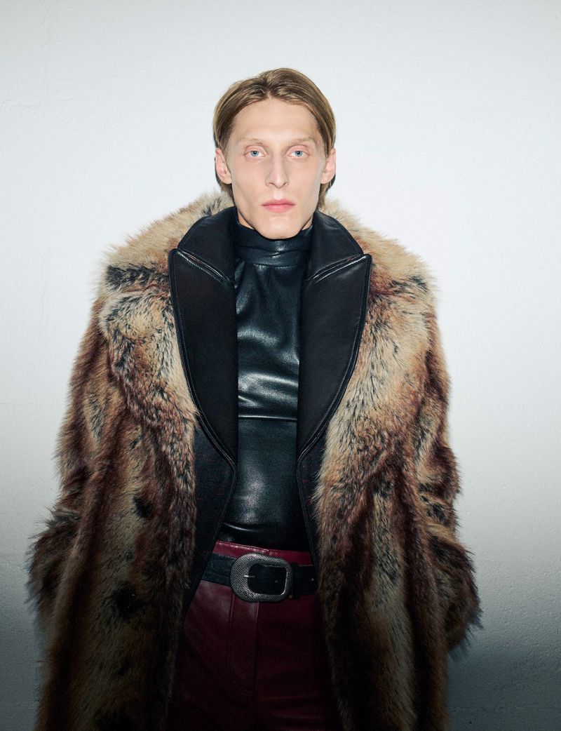 Elegant man in fur coat and leather top poses indoors. Fashion photography featuring a neutral color palette and soft lighting.