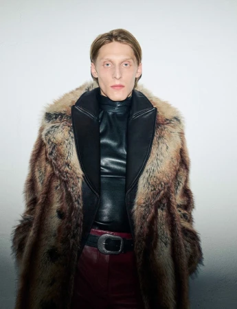 Elegant man in fur coat and leather top poses indoors. Fashion photography featuring a neutral color palette and soft lighting.
