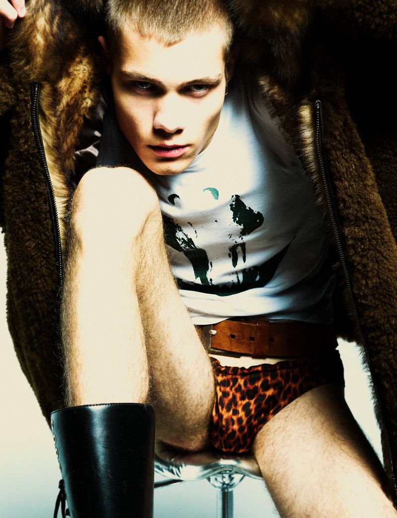 A fair-skinned man with light hair poses on a clear stool wearing a T-shirt, fur coat, leopard print underwear, and knee-high leather boots.