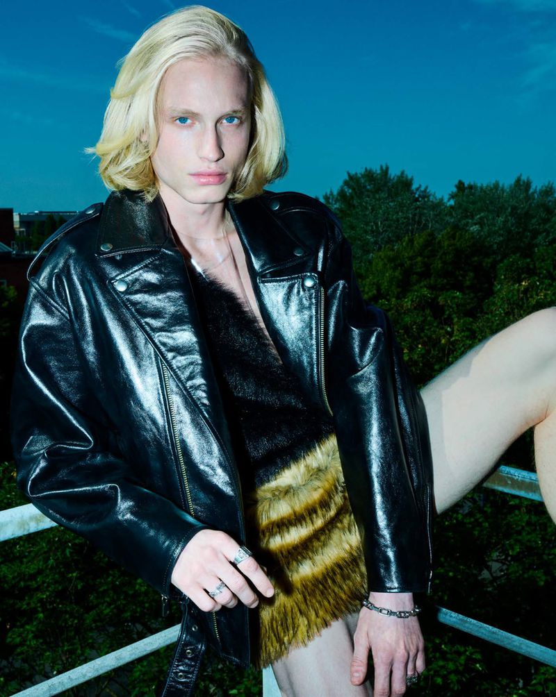 Blond male fashion model in leather jacket and fur top poses on rooftop with city trees and sky in background. Edgy style, urban scene.