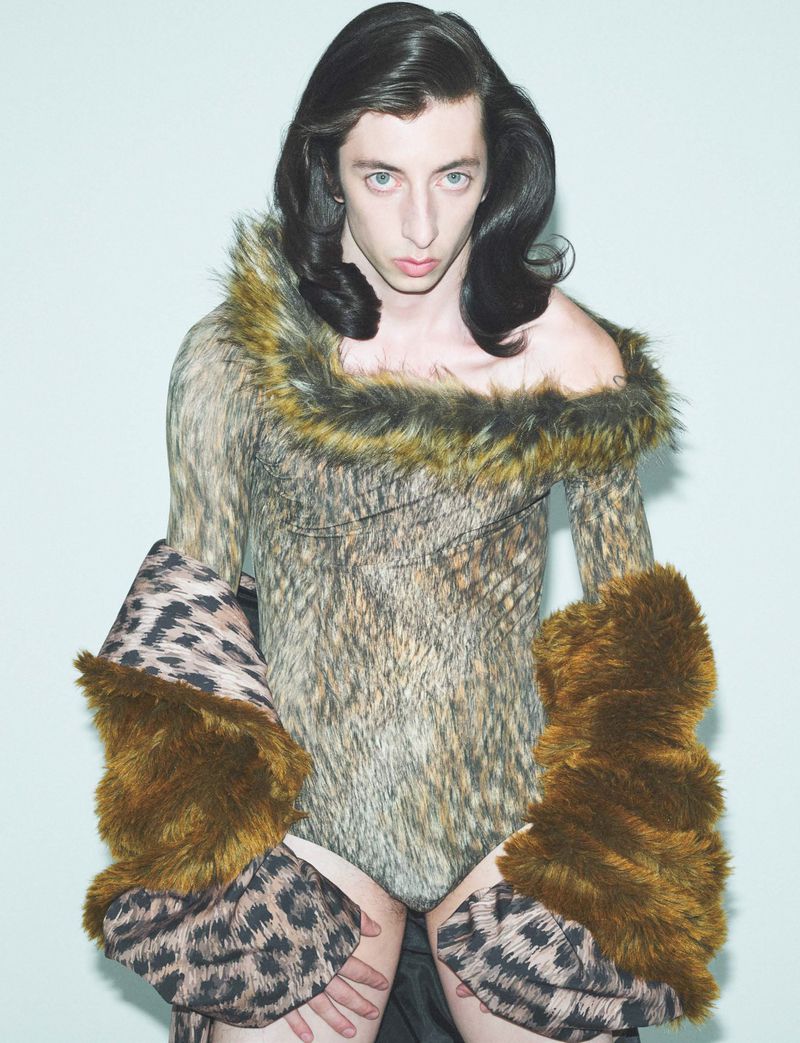 Adult posing in a studio wearing a fur-trimmed animal print bodysuit, leopard print sleeves with brown fur cuffs in front of a plain backdrop.