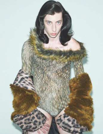 Adult posing in a studio wearing a fur-trimmed animal print bodysuit, leopard print sleeves with brown fur cuffs in front of a plain backdrop.