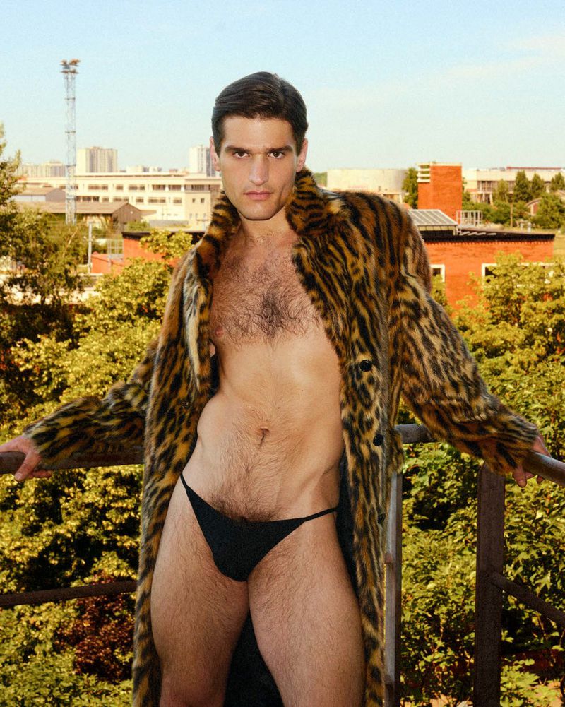 Adult male in black briefs and an animal-print coat poses on a balcony with a city skyline and trees in the background during daytime.