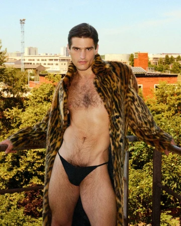 Adult male in black briefs and an animal-print coat poses on a balcony with a city skyline and trees in the background during daytime.
