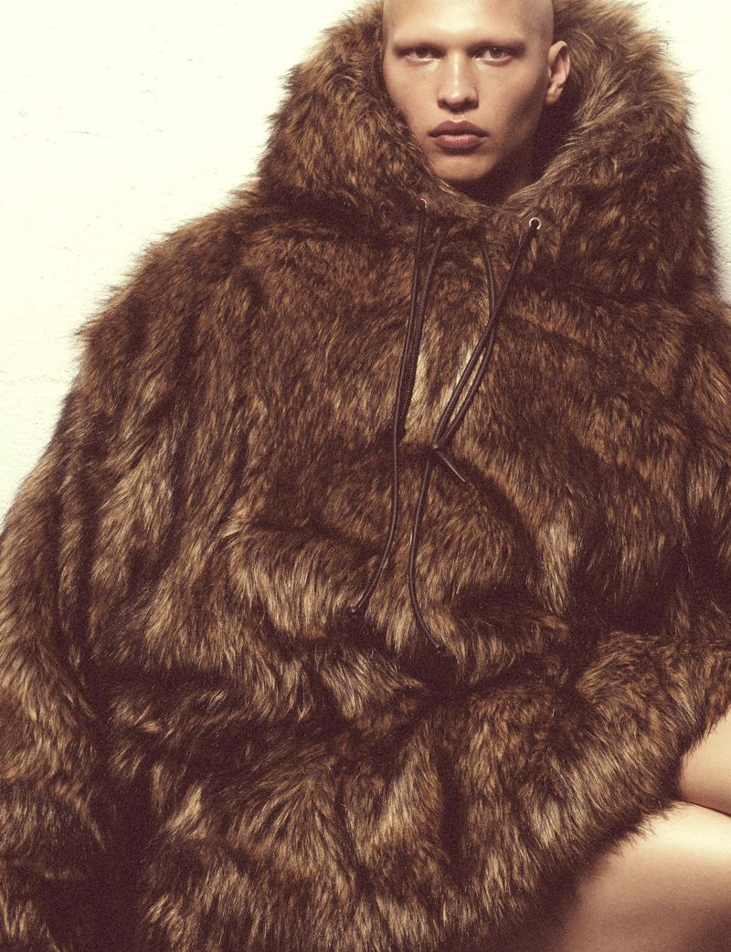 Bald adult male in a hooded poncho made of brown faux fur posing in front of a plain white wall in a studio-lit fashion shot.