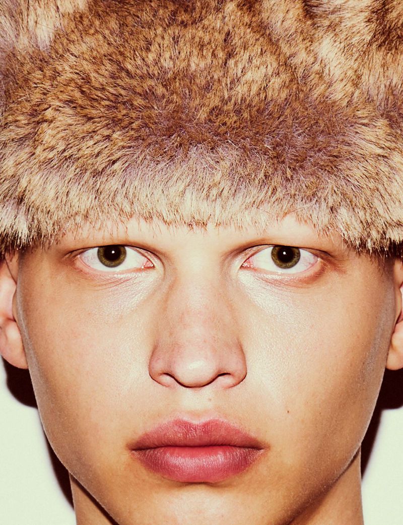 Close-up portrait of a fair-skinned person with light eyebrows wearing a fur hat, featuring a direct gaze and neutral facial expression.