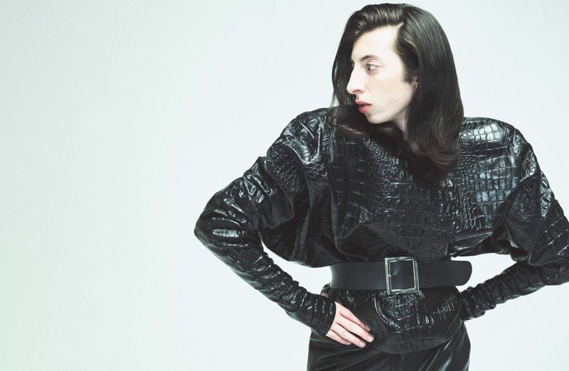 Woman in black faux crocodile jacket and wide belt poses against a white background, showcasing fashion and style.