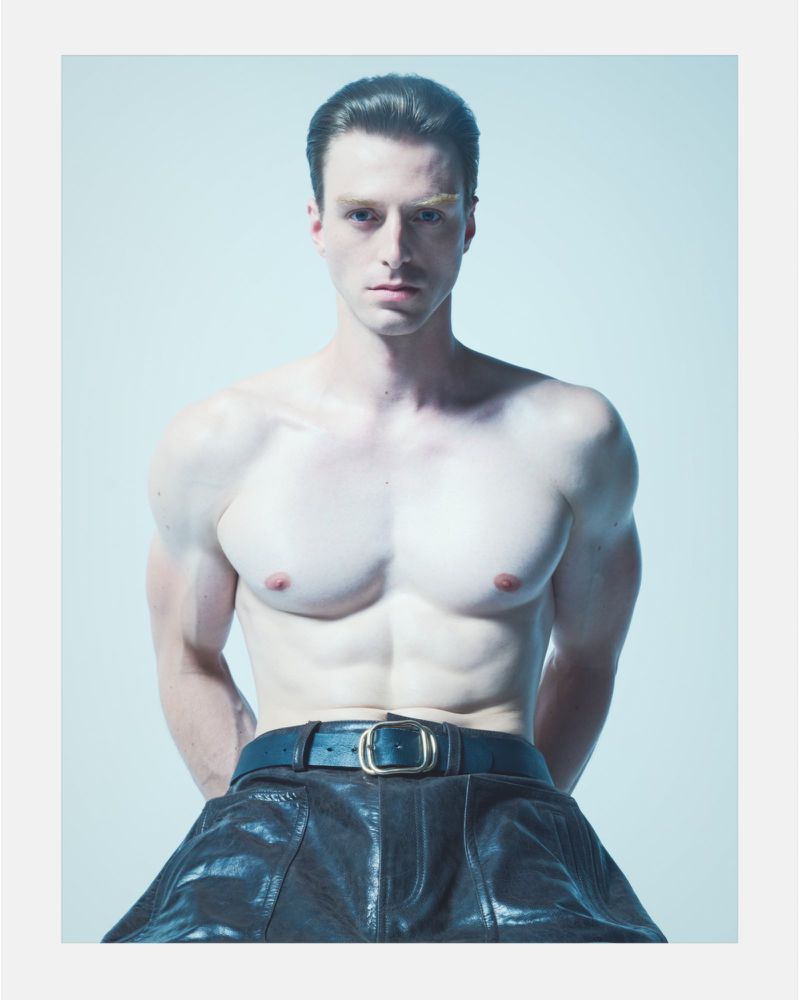 Portrait of a muscular man with slicked-back hair wearing leather pants and a gold belt buckle, posing against a light blue backdrop.