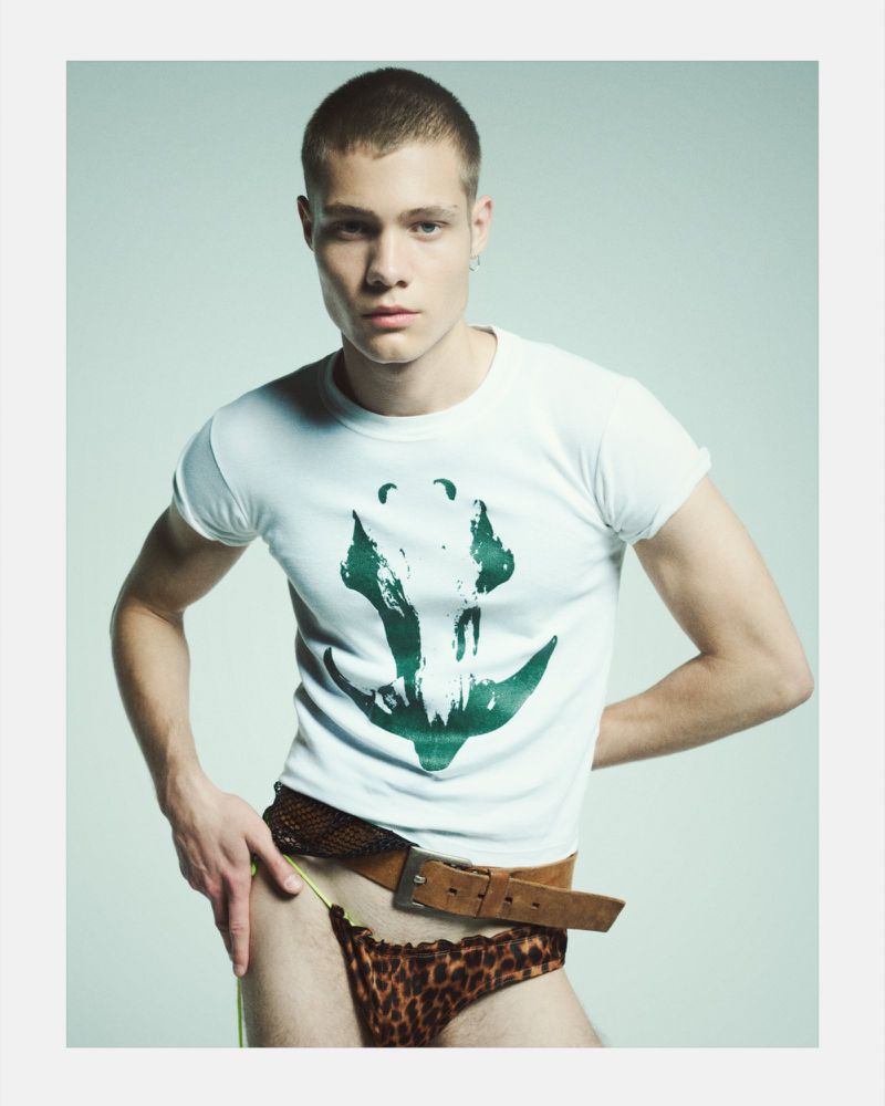 Young adult man in studio setting wearing a white tee shirt with a green print design, leopard print underwear and a brown belt.