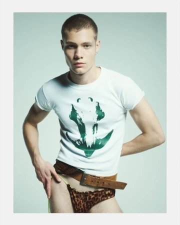 Young adult man in studio setting wearing a white tee shirt with a green print design, leopard print underwear and a brown belt.