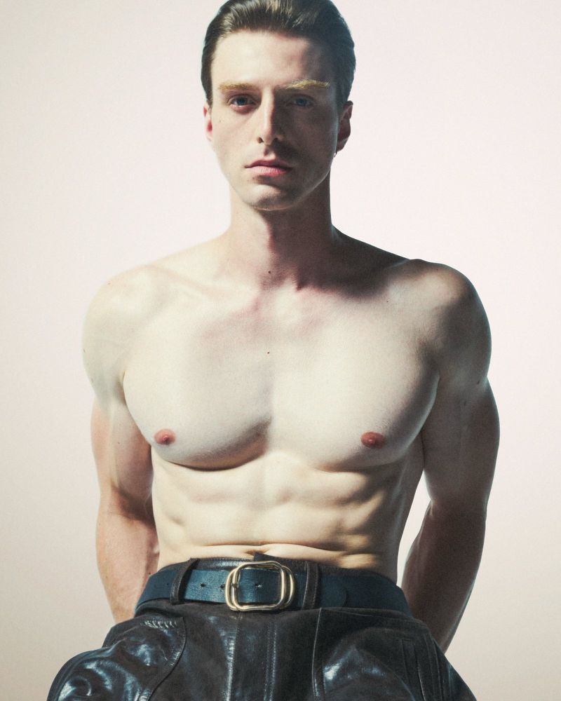 Shirtless man posing in leather pants with muscular physique against a neutral backdrop in a studio photography setting.