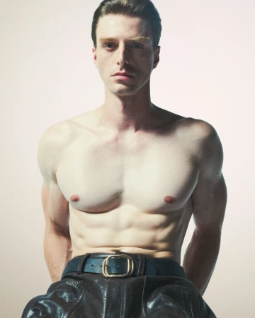 Shirtless man posing in leather pants with muscular physique against a neutral backdrop in a studio photography setting.