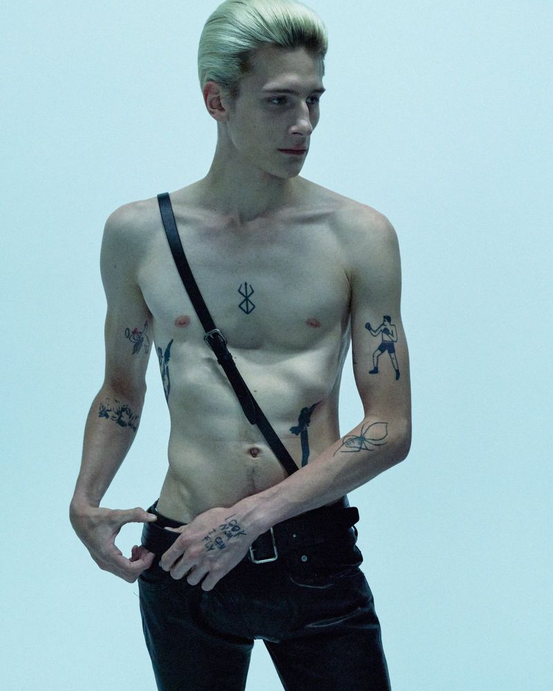 Tattooed male fashion model with platinum blonde hair, shirtless, wearing black leather pants and suspenders. Standing against a plain light blue background.