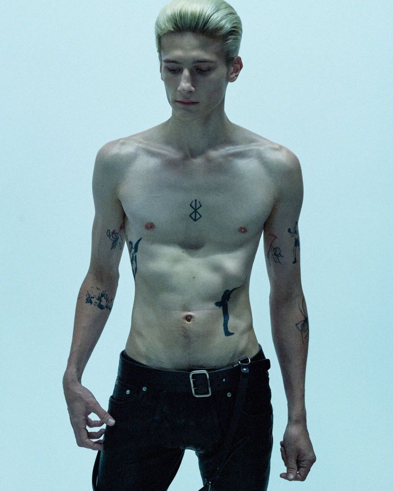 Shirtless young man with slicked-back blonde hair and torso tattoos wearing black pants against a pale studio backdrop.
