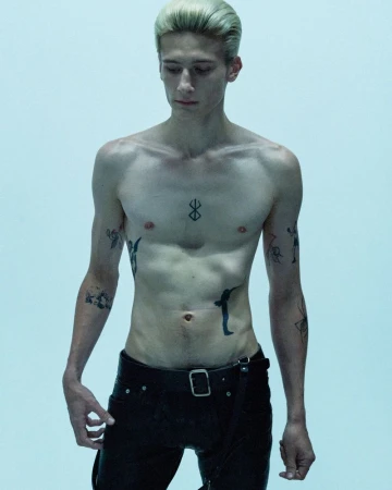 Shirtless young man with slicked-back blonde hair and torso tattoos wearing black pants against a pale studio backdrop.