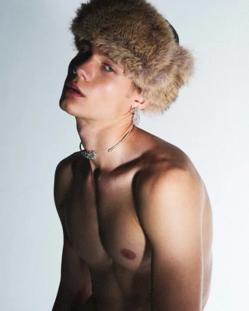 Headshot of a shirtless fair-skinned male model wearing a fur hat and choker against a white backdrop in a portrait-style composition.