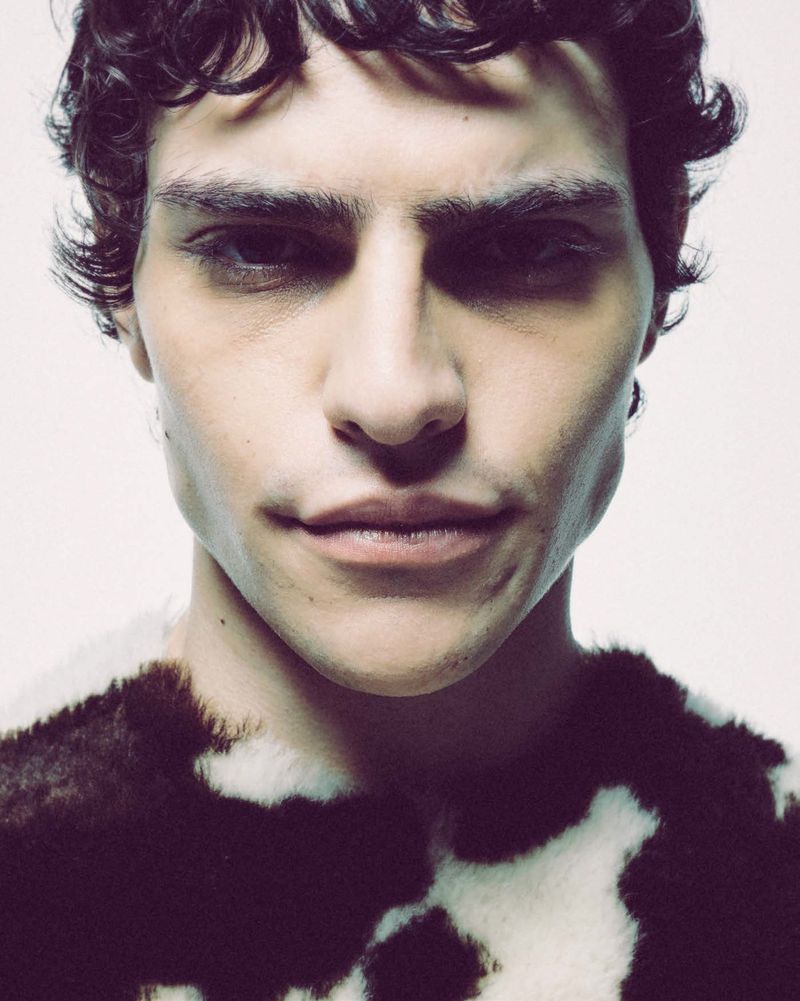 Close-up portrait of a young adult male with curly hair wearing a patterned top, against a plain white backdrop in a photo studio.