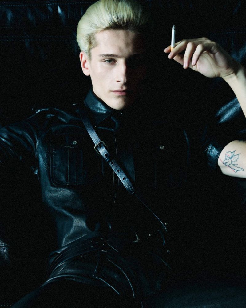 Studio portrait of a fair-skinned adult male with blond hair, wearing a black leather outfit, holding a cigarette, and sitting in a dark setting.