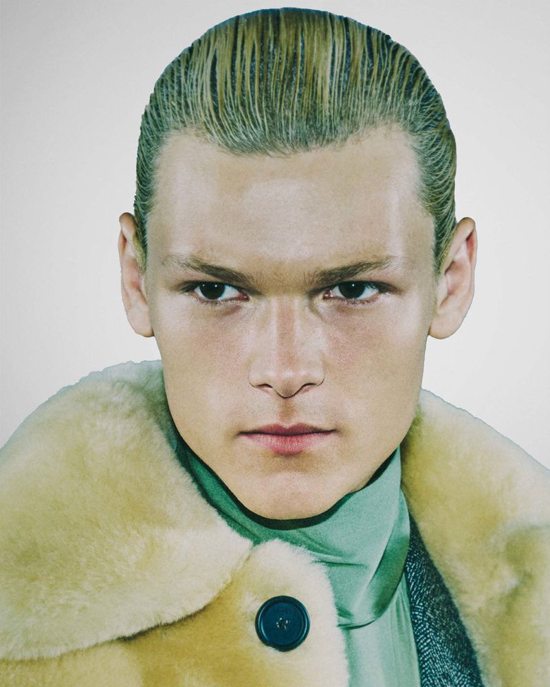 Close-up studio portrait of a young adult with slicked-back blonde hair, wearing a fuzzy tan coat and teal turtleneck.