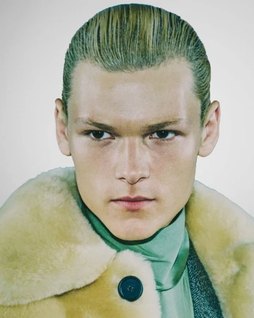 Close-up studio portrait of a young adult with slicked-back blonde hair, wearing a fuzzy tan coat and teal turtleneck.