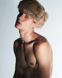 Shirtless young man wearing a brown fur ushanka hat and silver choker necklace against a light grey background