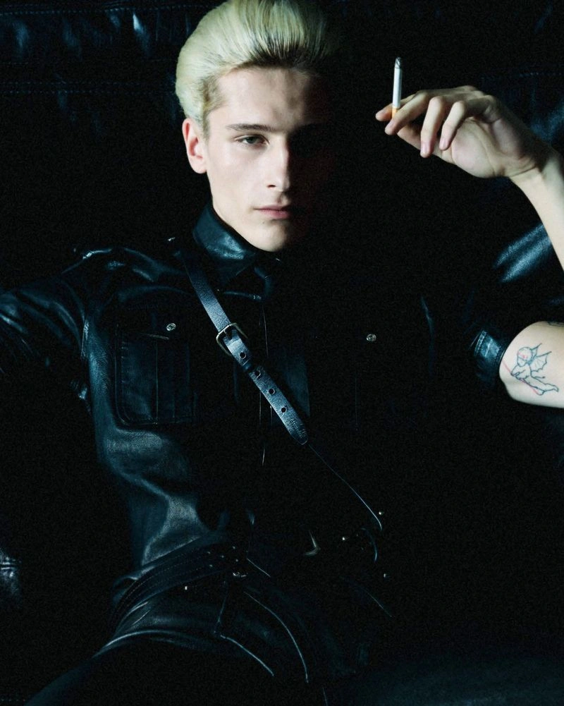 Blonde male model wearing a black leather shirt and shoulder harness, holding a cigarette against a dark background.
