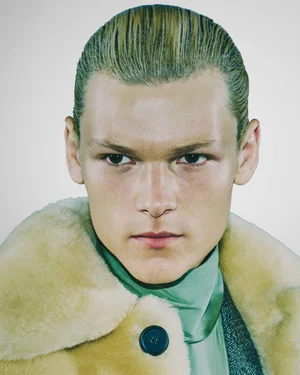 Close-up portrait of a male model with slicked-back blonde hair wearing a green turtleneck and cream shearling coat against a plain background.