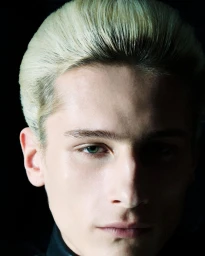 Close-up portrait of a young man with slicked-back platinum blonde hair lit in chiaroscuro against a black background.