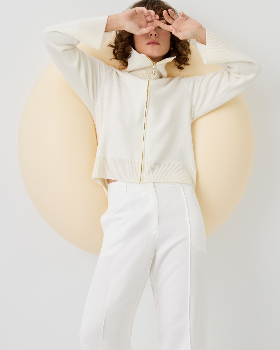 Woman in white zip-up sweater and pants poses in front of an off-white circular shape against a bright white background in a studio shot.