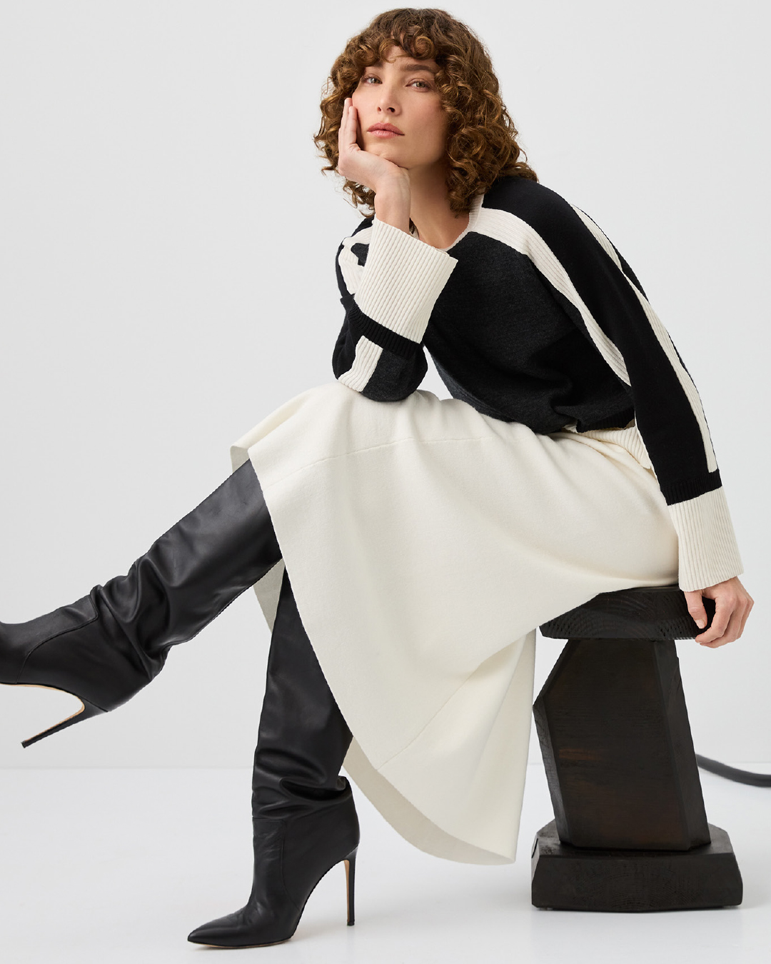 A woman with curly hair in a black and white sweater and cream skirt poses sitting on a stool, wearing black heeled boots against white.