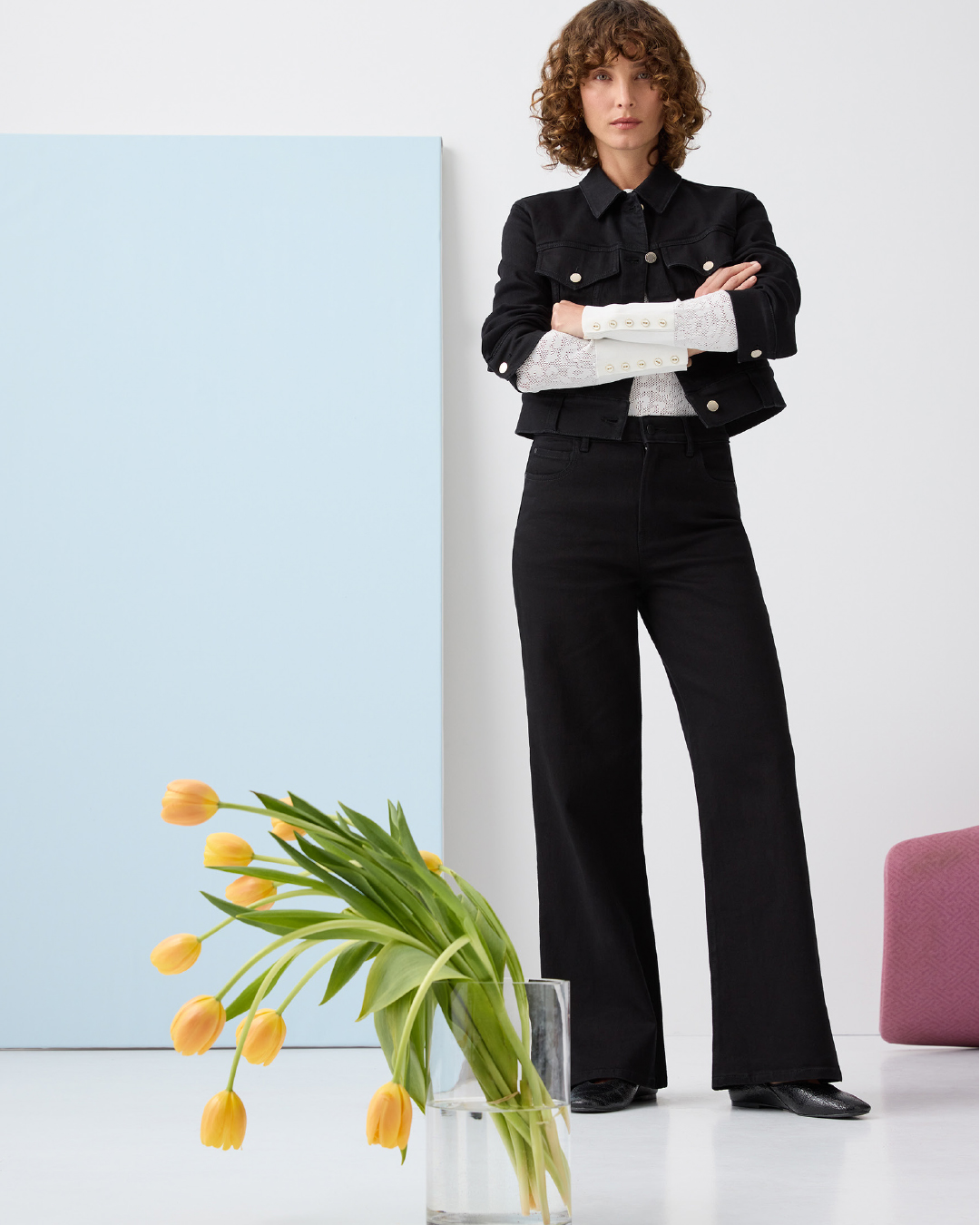 A woman in a black denim jacket and black pants stands in a photo studio with a vase of yellow tulips next to her.