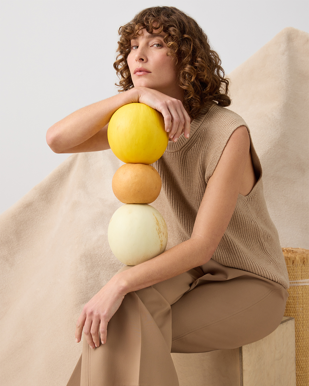 Woman in tan outfit sitting with stacked melons in studio, showcasing fashion and healthy eating with soft lighting and neutral tones.
