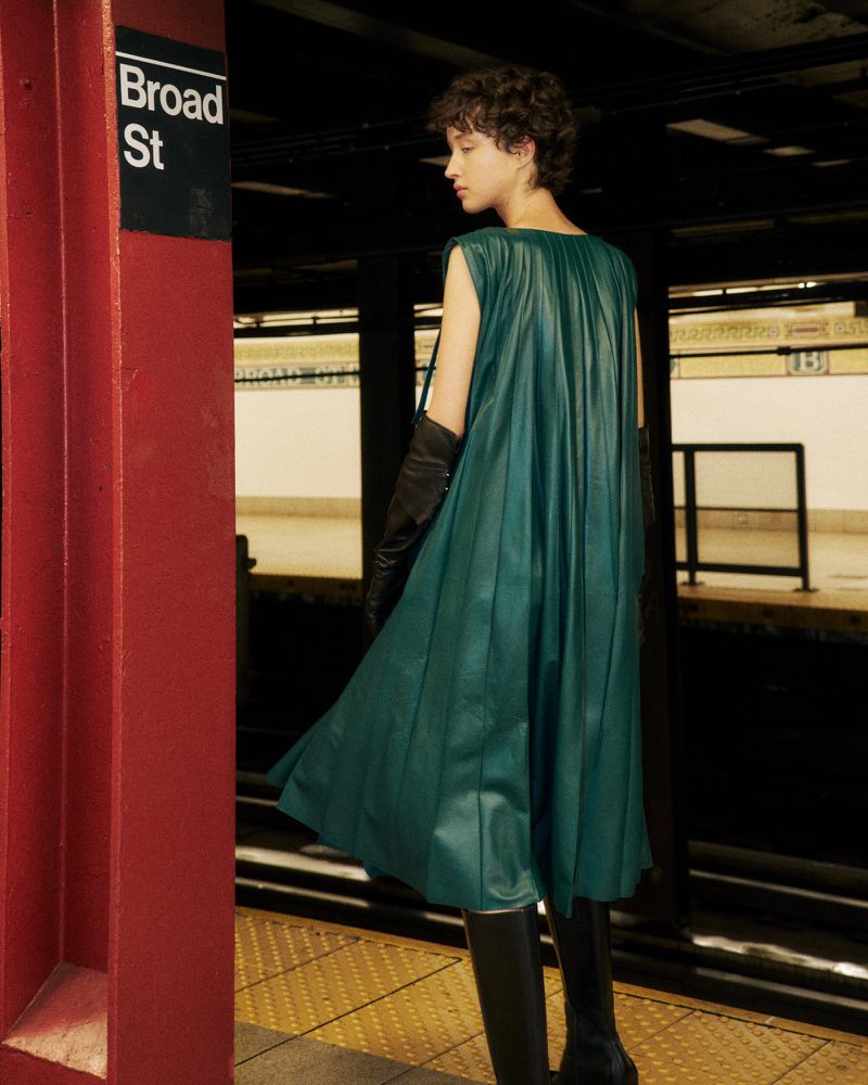 A slender adult fashion model in a green dress, long black gloves, and boots stands on a subway platform near the Broad St sign.