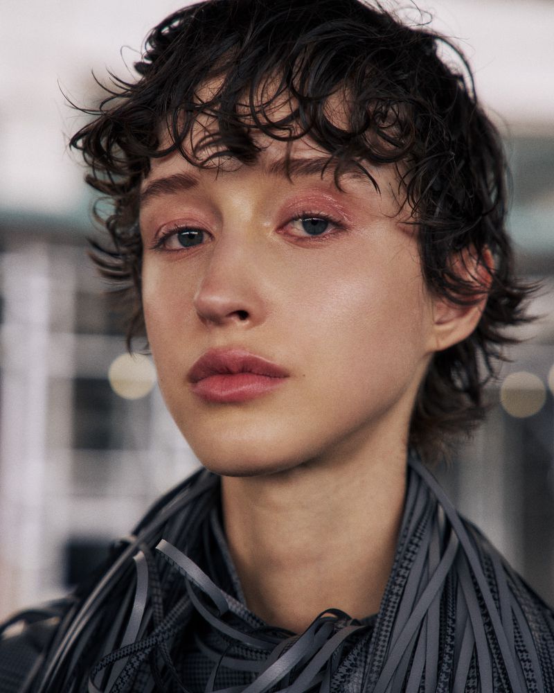 Close-up portrait of a young adult woman with short, curly dark hair, wearing a dark-colored textural garment, indoors with blurred background.