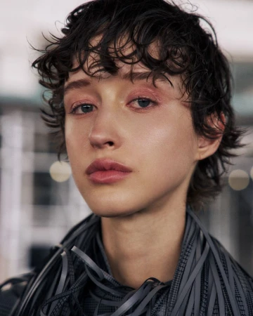 Close-up portrait of a young adult woman with short, curly dark hair, wearing a dark-colored textural garment, indoors with blurred background.