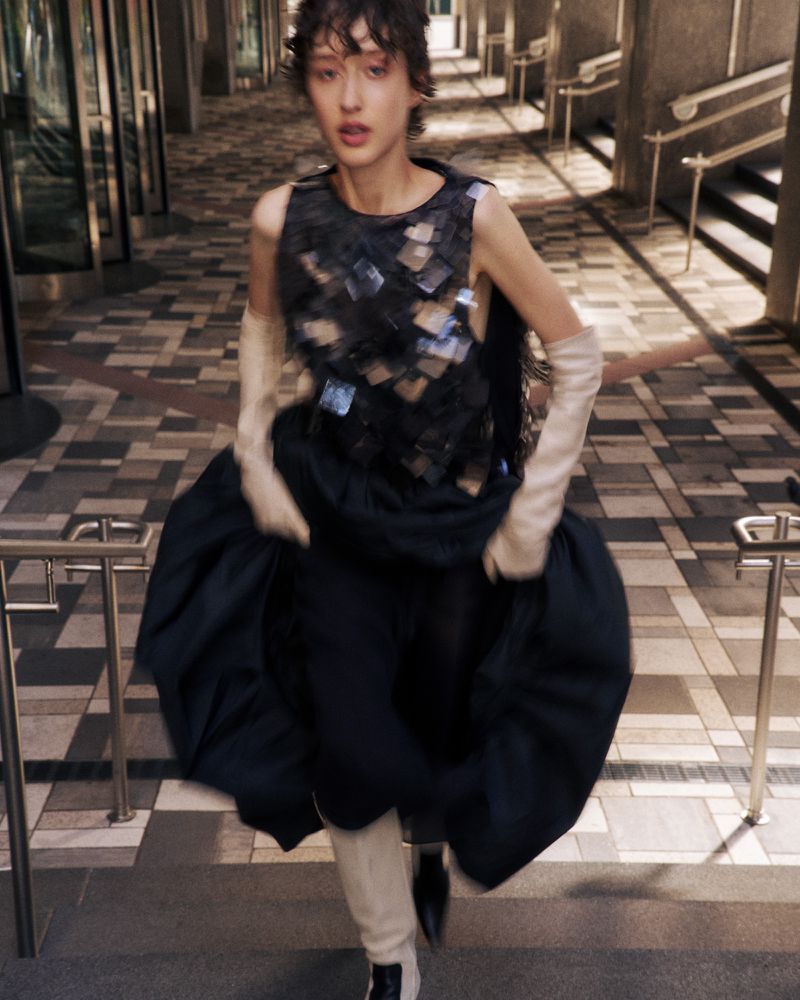 Fashion photography of a woman in a dramatic black dress with a silver pattern, long white gloves, and dark boots, running through a hallway.