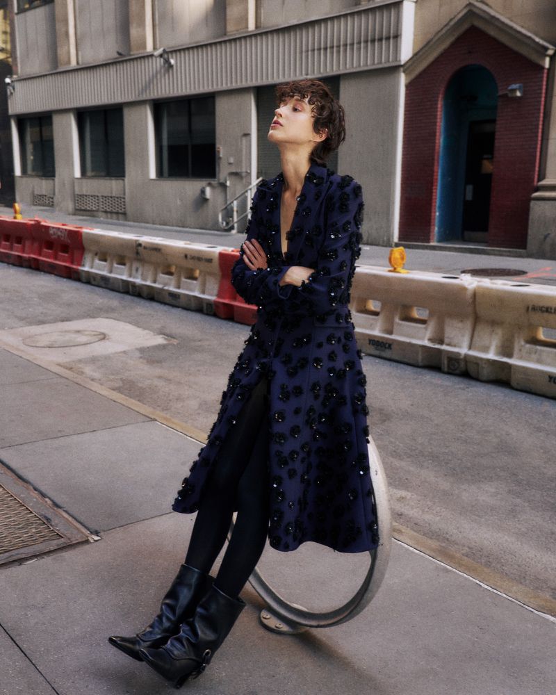 Woman in navy floral coat, black tights, and boots poses on an urban street with a building in the background on a sunny day.