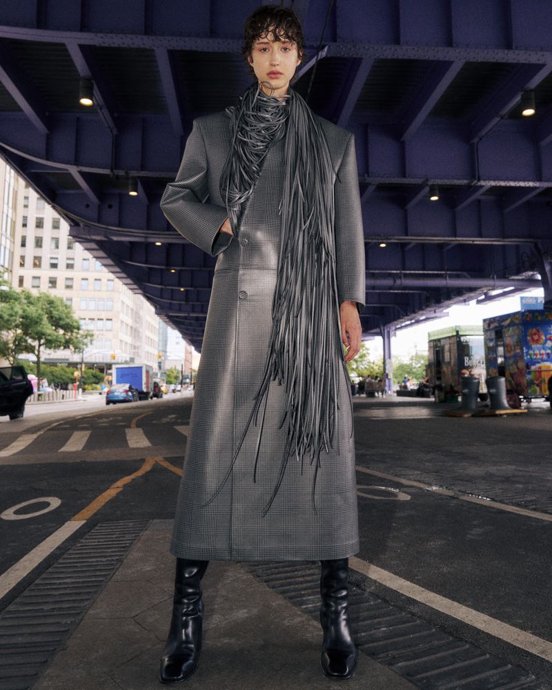 A fashion-forward woman in a gray coat and black boots stands confidently on a city street under a bridge, showcasing modern style.