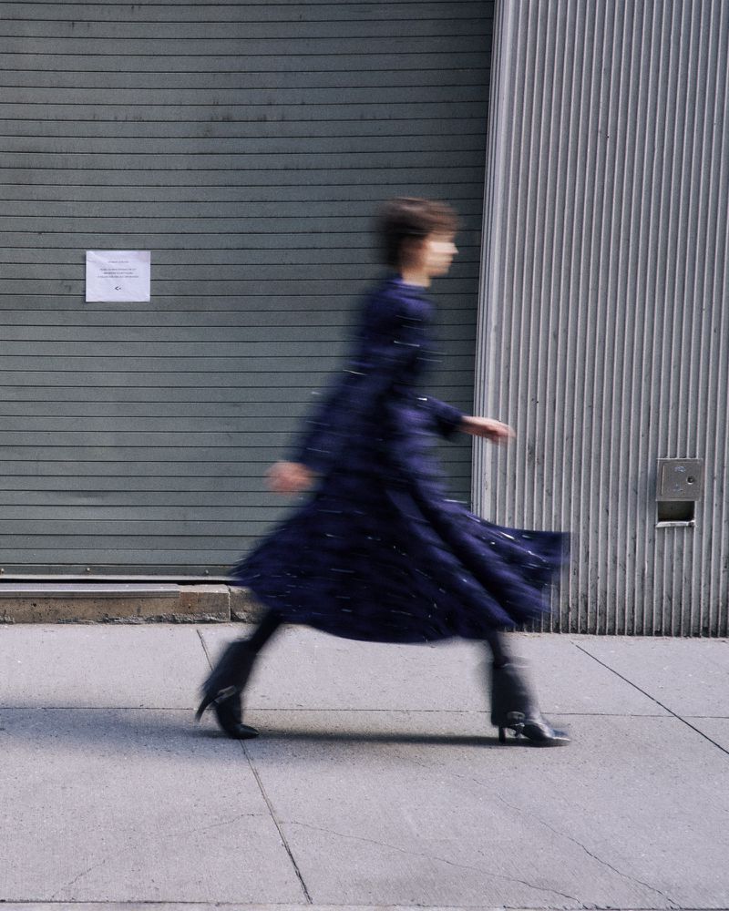 Blurred motion shot of a woman in a dark blue dress and boots walking on a city sidewalk with a corrugated metal wall background.
