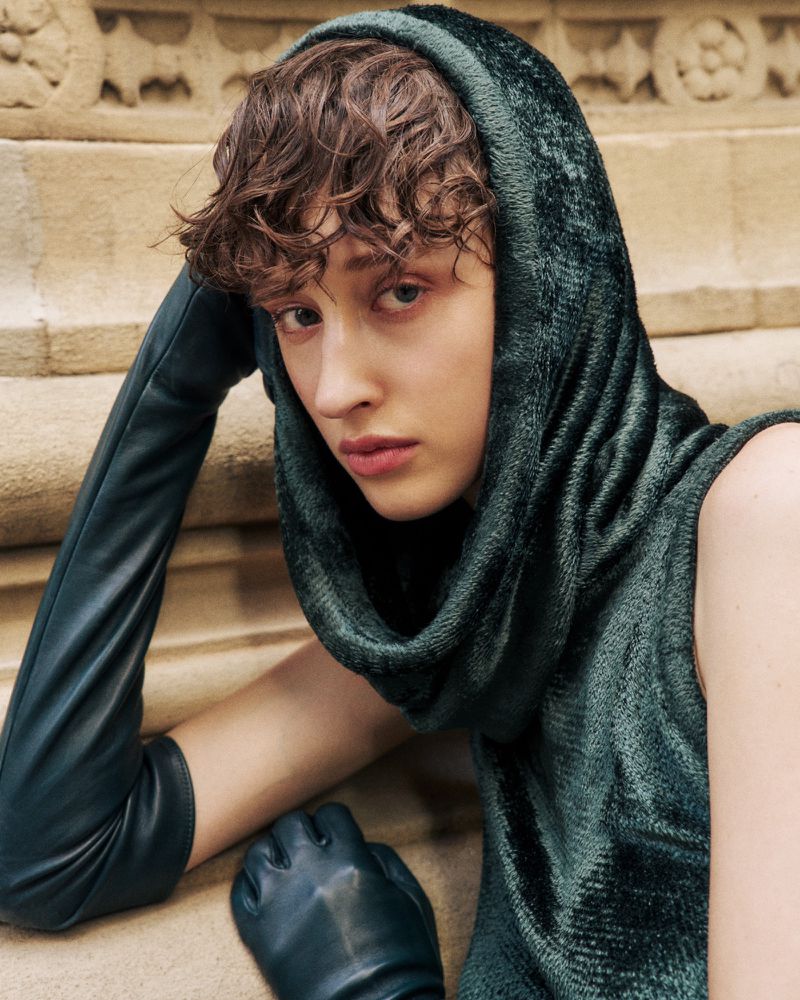 Close-up studio shot of a young adult wearing a hooded, sleeveless, dark green velvet top and dark green leather opera gloves, staring at the camera.