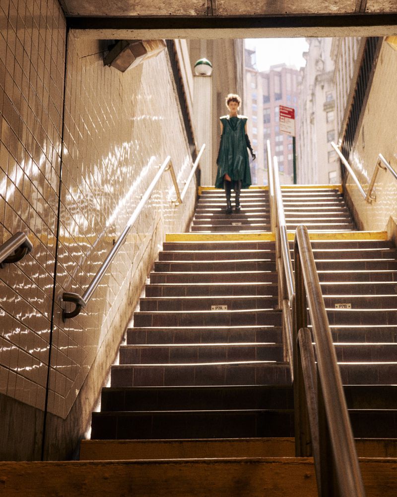Woman in a teal dress ascends a long staircase from a subway station into the city street, bathed in warm, directional sunlight.